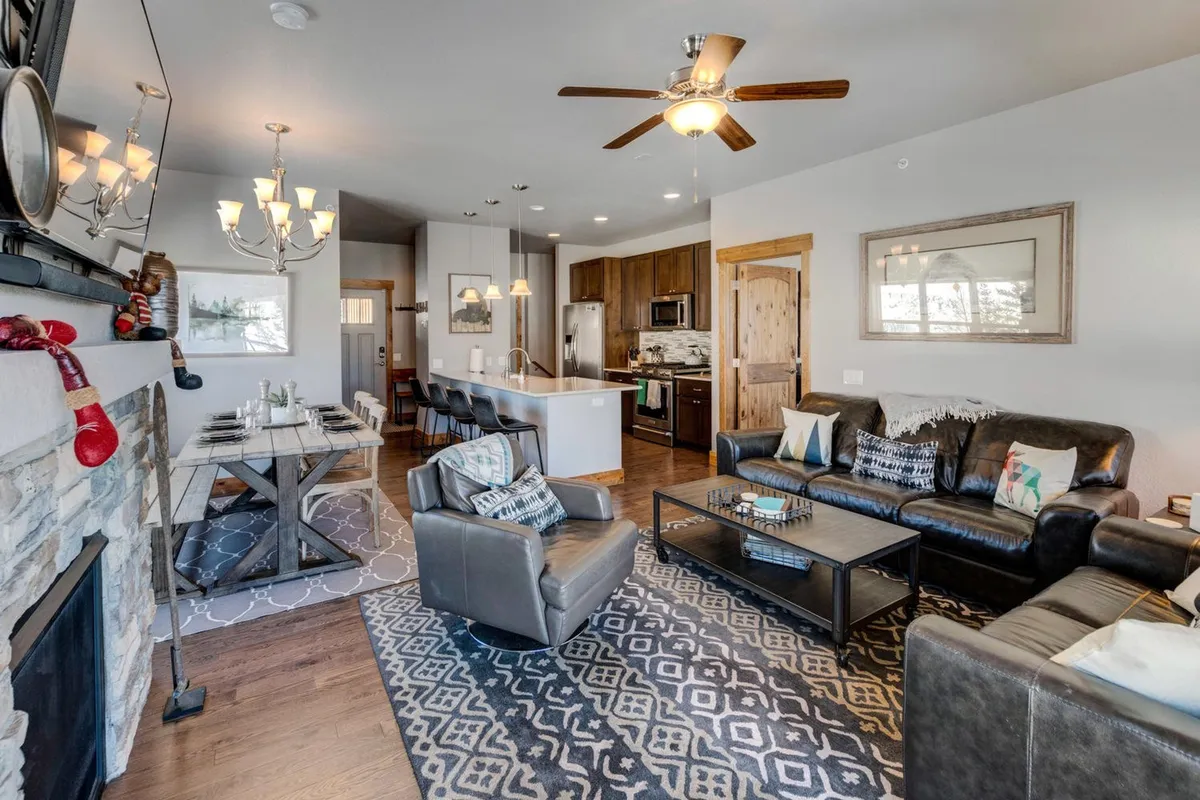 A spacious open-concept living and dining area featuring a cozy fireplace, a generous dining table set for a meal, plush living room seating, and a modern kitchen island in the background. Sunlight streams in from large windows, creating a bright and inviting space perfect for gatherings.