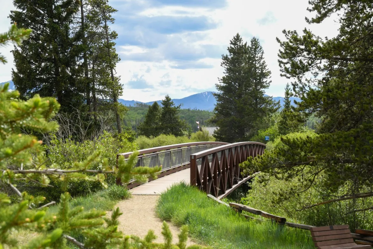 A tranquil exterior scene showing a wooden bridge set amidst rich greenery, with a dramatic mountain backdrop. The environment invites walking and hiking, representing easy access to nearby outdoor trails.