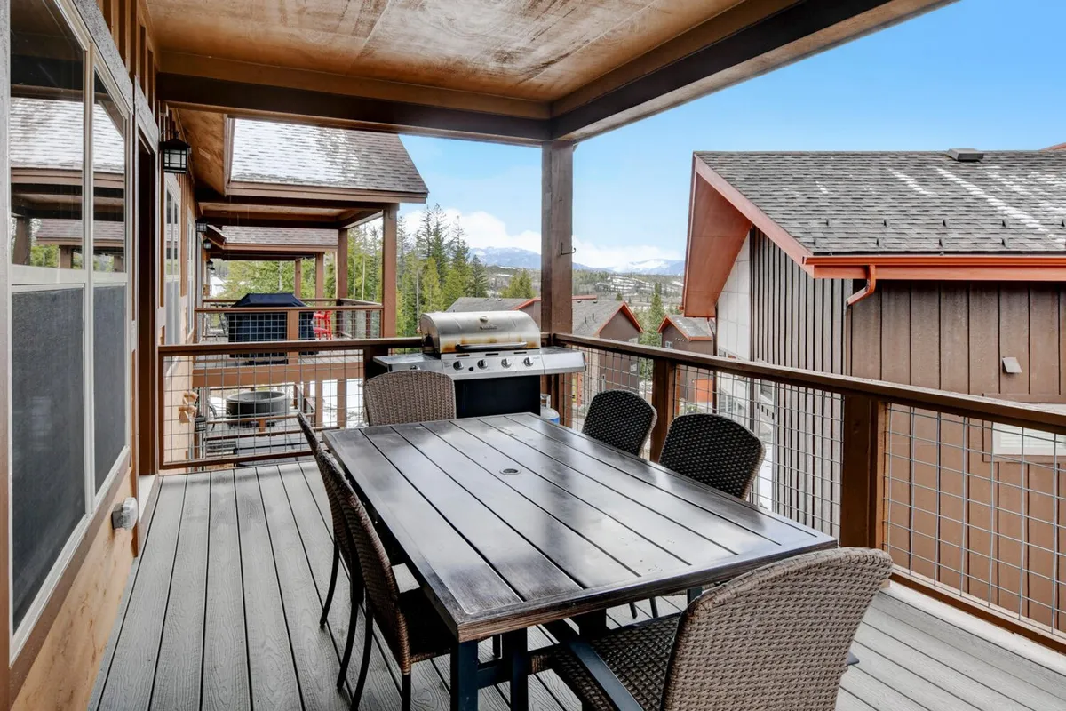 A relaxed outdoor dining area on a patio with a table set for a meal, a nearby grill, and scenic views of surrounding trees and mountains. The space is designed for al fresco dining, family barbecues, and making the most of warm weather.