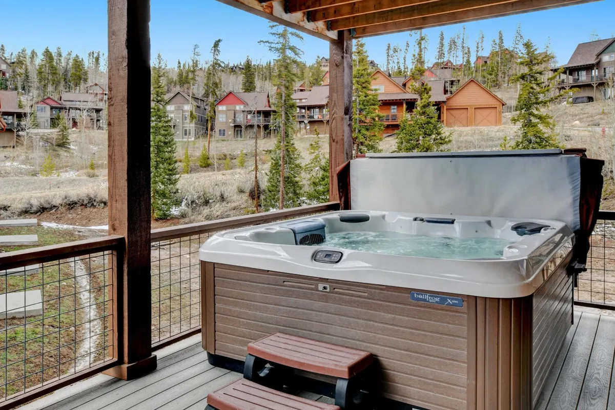A scenic exterior view featuring a private hot tub situated on a wooden deck, surrounded by lush landscape and mountain views. The area is set up for relaxation and social gatherings, with the setting sun casting a warm glow and offering the opportunity for stargazing at night.