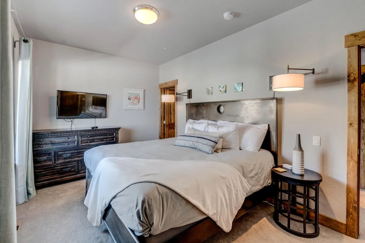 A tranquil king bedroom featuring a plush bed dressed in premium linens, soft sunlight streaming through the windows, tasteful wall art, and a television atop the dresser. The scene exudes comfort and serenity, suggesting a luxurious retreat ideal for restful nights.