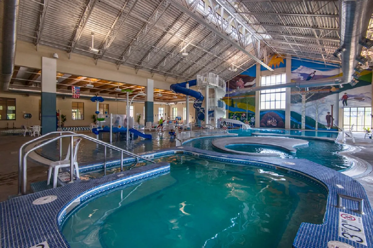 An energetic indoor recreation area with a bright pool, a colorful water slide, and a vibrant mural depicting adventure, where children are playing in the water as parents relax nearby.