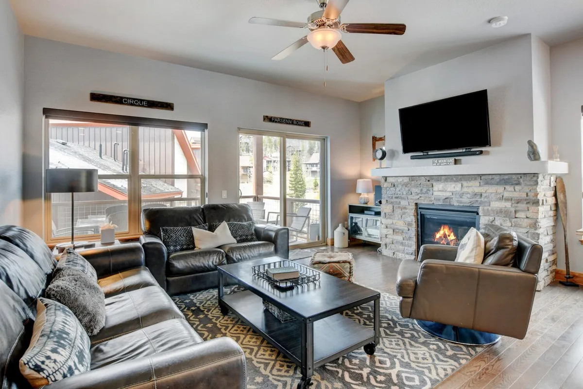 A cozy living room with a stone fireplace, deep and comfortable seating, large windows offering stunning outdoor views, and tasteful mountain-inspired decor. The room invites relaxation and togetherness, with soft natural light and a welcoming ambiance.