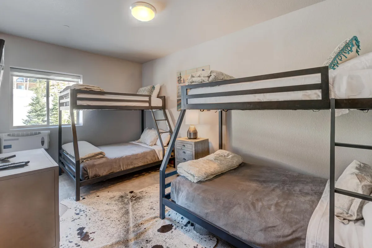 A playful bunk room with twin and full beds featuring colorful bedding, a nightstand with reading lamp, and a window with scenic outdoor views. The atmosphere is lighthearted and inviting—perfect for kids or friends to unwind and create memories.