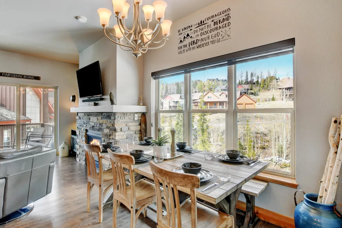 An elegant dining area anchored by a large, rustic wooden table, illuminated by a striking chandelier. A fireplace adds warmth, and a wall of windows frames picturesque mountain views, bathing the room in natural light and emphasizing the mountain setting.