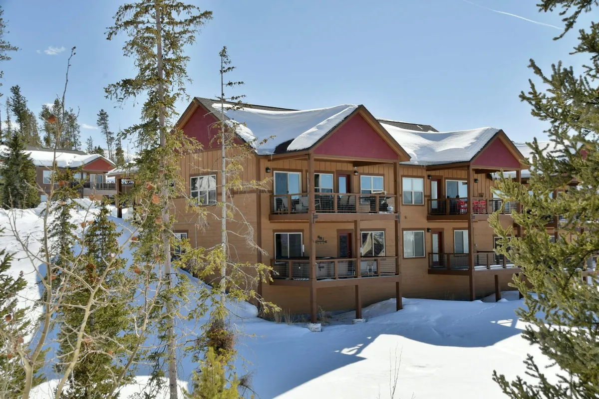 The image showcases a beautiful mountain villa surrounded by snow-covered ground and tall pine trees. The home's inviting wooden balconies overlook a serene winter landscape, with architectural details blending luxury and alpine charm. The setting suggests opportunities for skiing, snowboarding, and relaxing outdoors, enhanced by the tranquil ambiance of fresh snowfall.